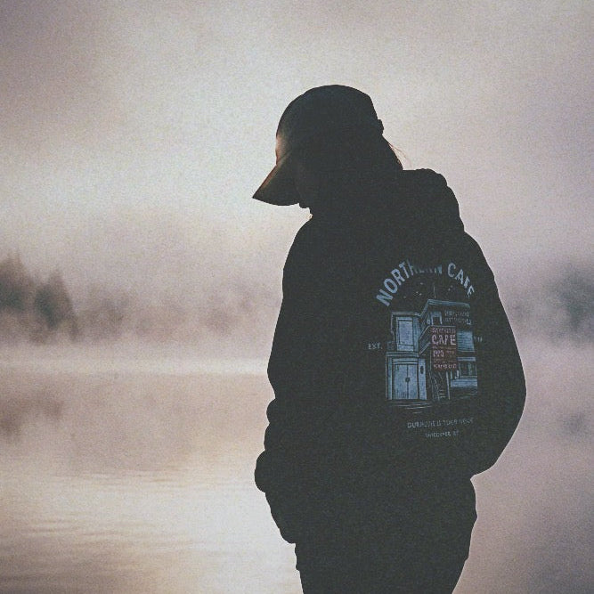 Person wearing a hoodie with a visible brand logo standing by a misty lake.
