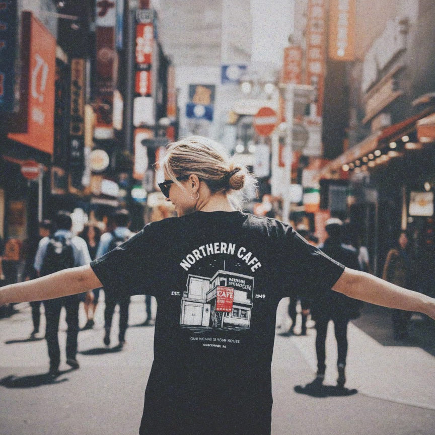 Person wearing a black t-shirt with 'Northern Cafe' logo in a busy city street.