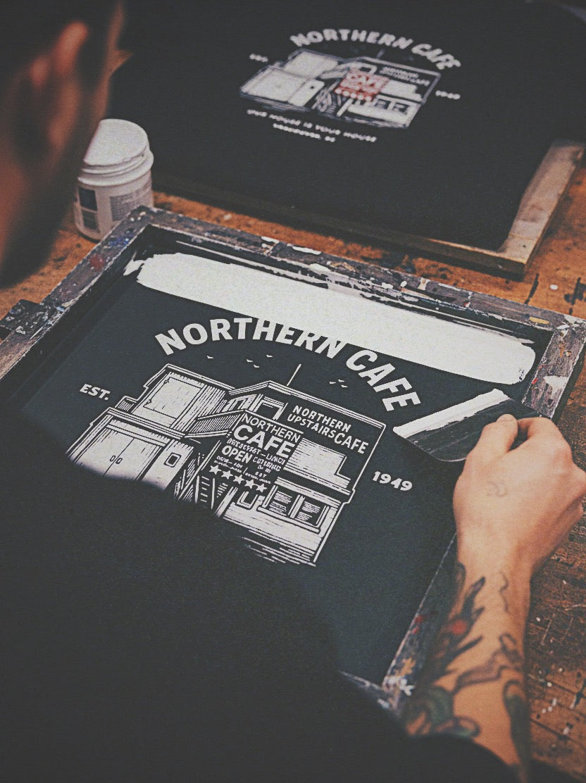 Person working on a print of 'Northern Cafe' with tattooed hands.