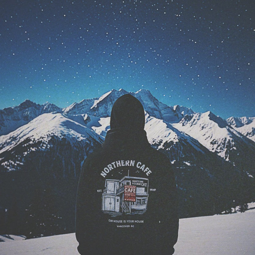 Person wearing a hoodie with 'Northern Cafe' design standing in front of snow-covered mountains under a starry night sky.