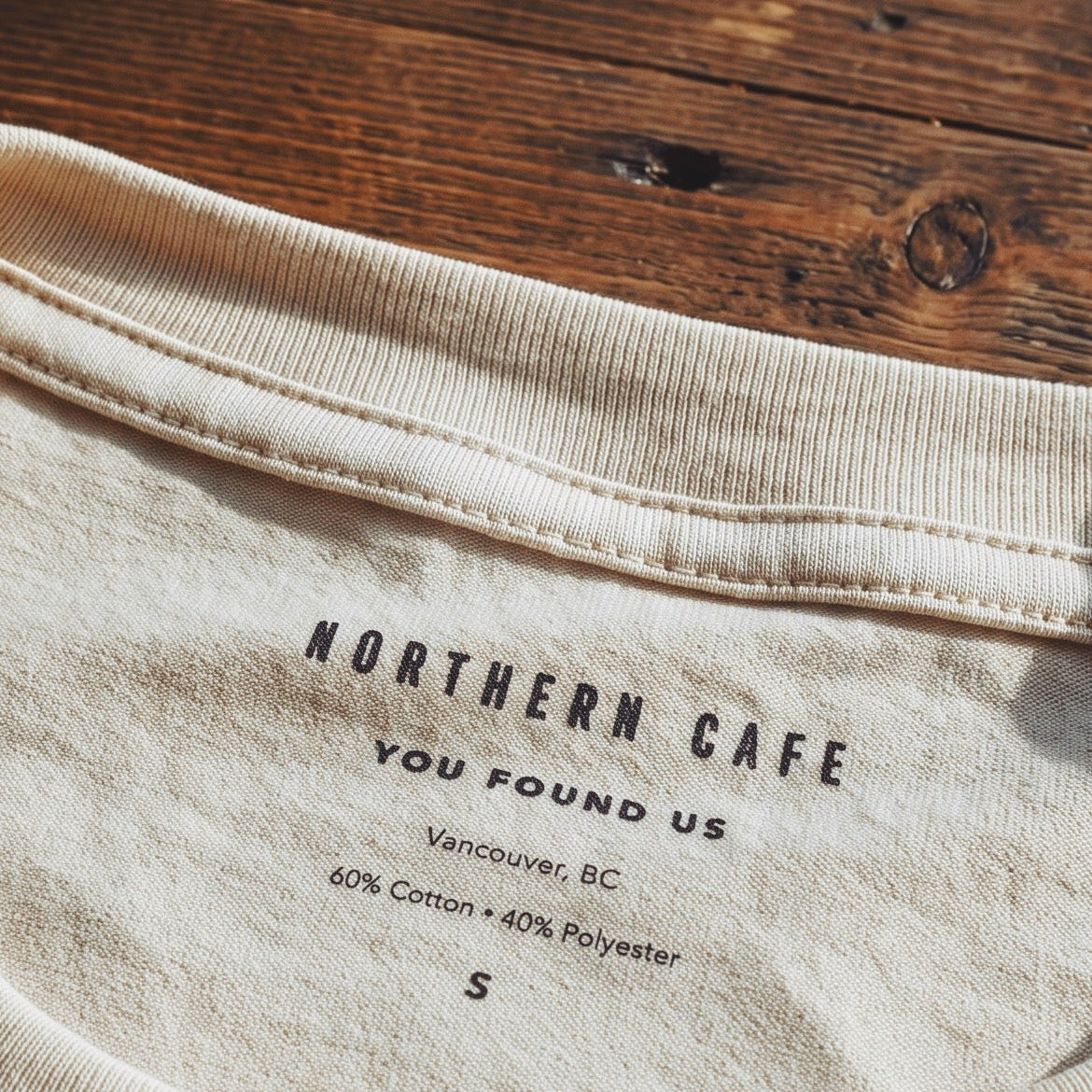 Close-up of a cream-colored t-shirt collar laid flat on a dark wooden surface, showing the printed neck label text 'NORTHERN CAFE YOU FOUND US'.