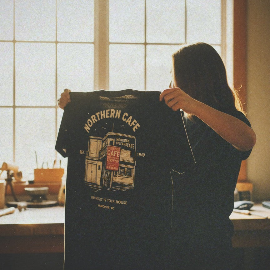 Person holding a t-shirt with 'Northern Cafe' design in a room with large windows.
