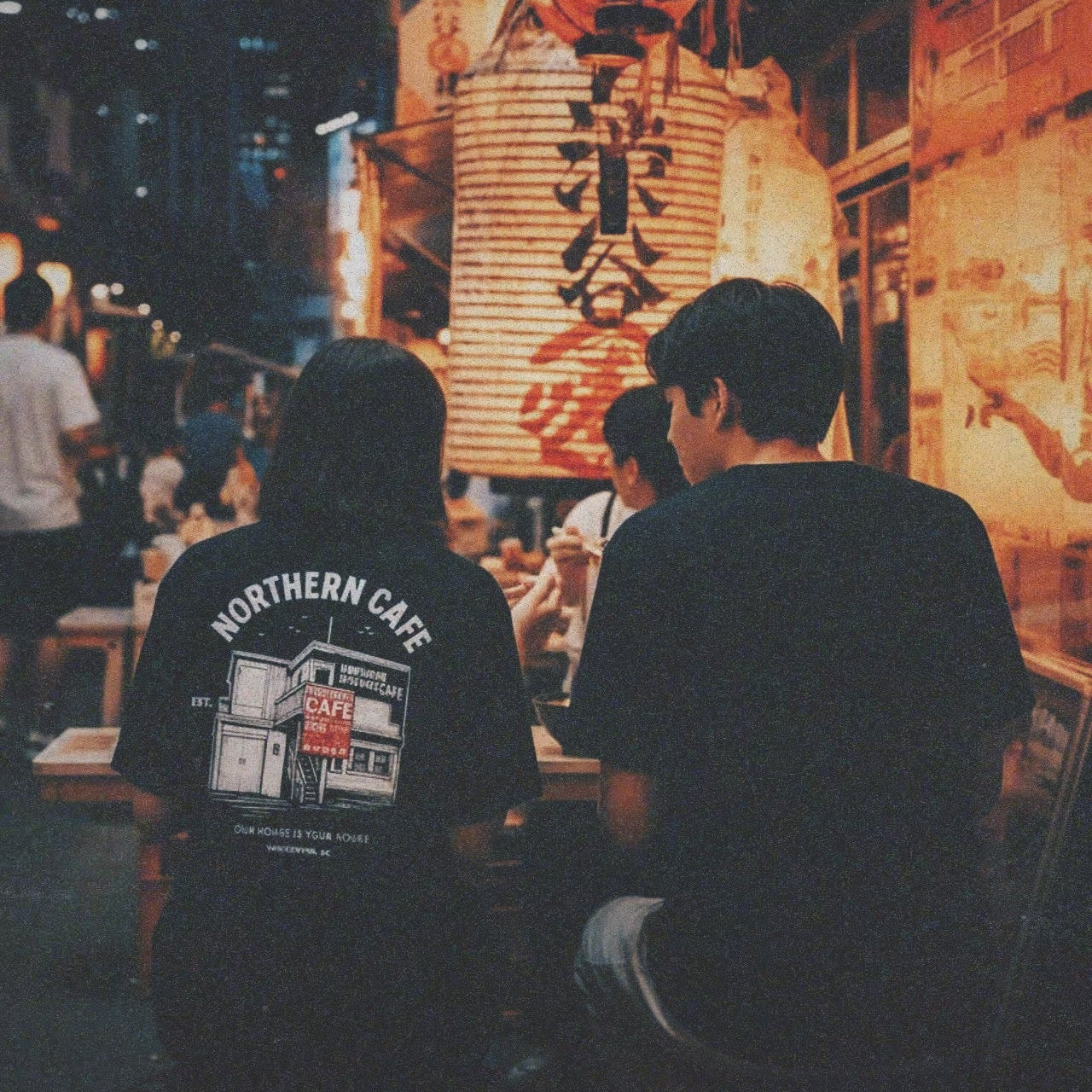 Two people sitting at a table in an urban setting with neon signs and city lights.