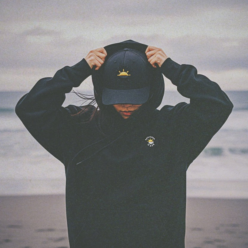 Person wearing a black hoodie with a logo, standing on a beach with ocean in the background