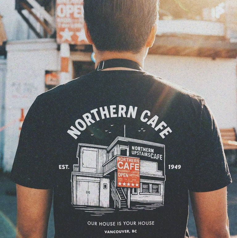 Person wearing a black t-shirt with 'Northern Cafe' design, standing in front of a cafe.