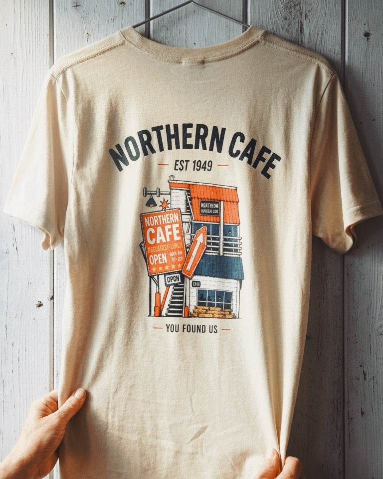 A beige graphic t-shirt hanging on a wooden hanger against a gray wood-paneled wall, with hands holding the bottom corners of the shirt.