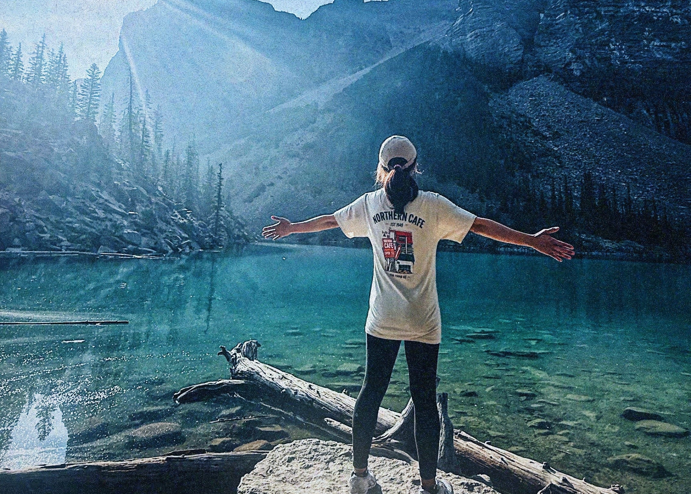 A person stands on a large rock by a turquoise mountain lake with arms outstretched, wearing a white graphic T-shirt, black leggings, and a light-colored cap, with rocky mountains and a blue sky in the background.