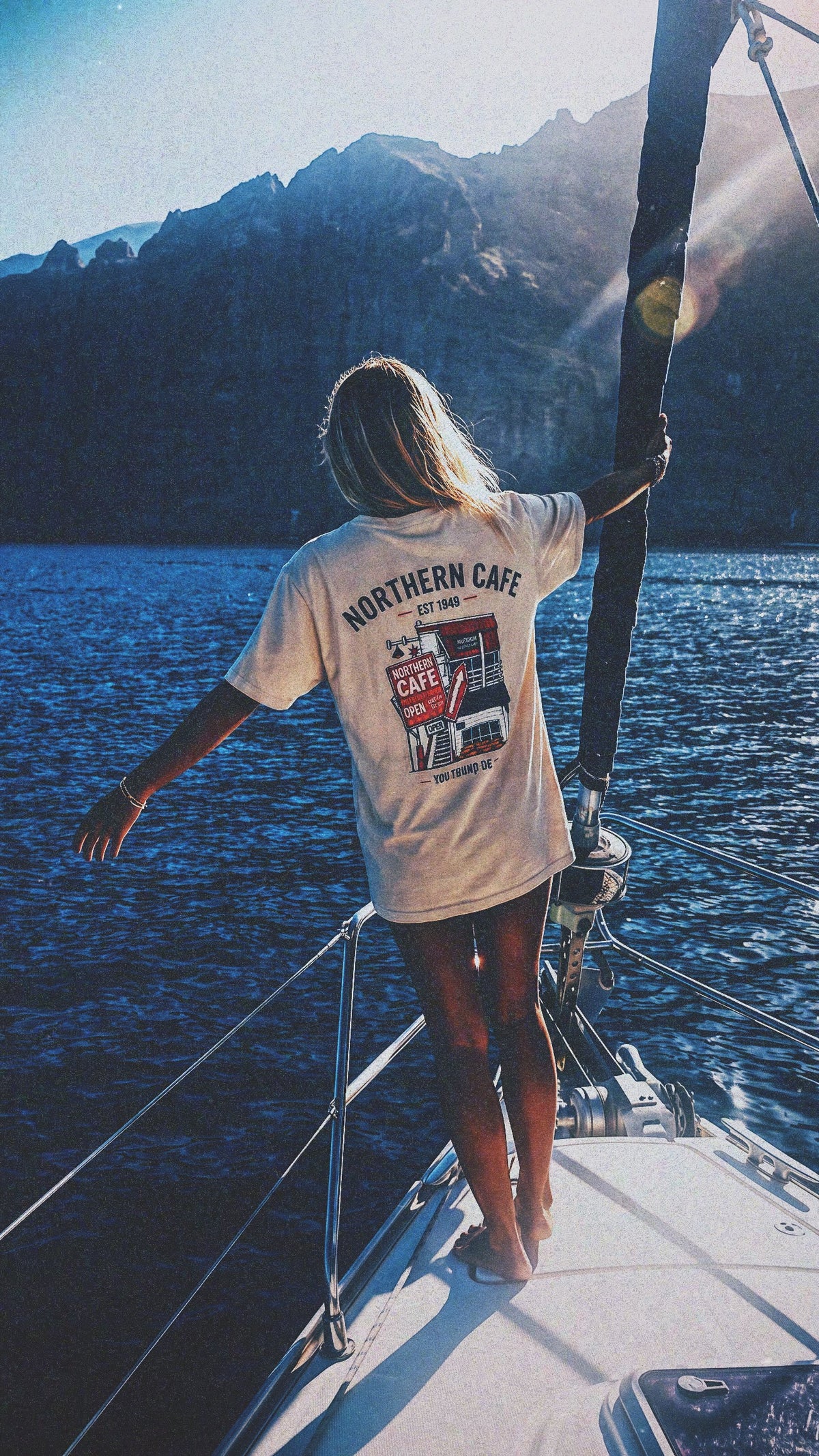 A woman stands on the bow of a sailboat, seen from behind, wearing a white graphic t-shirt with 'NORTHERN CAFE' printed on the back. The background features a scenic view of deep blue water and dark mountains under a bright sun.